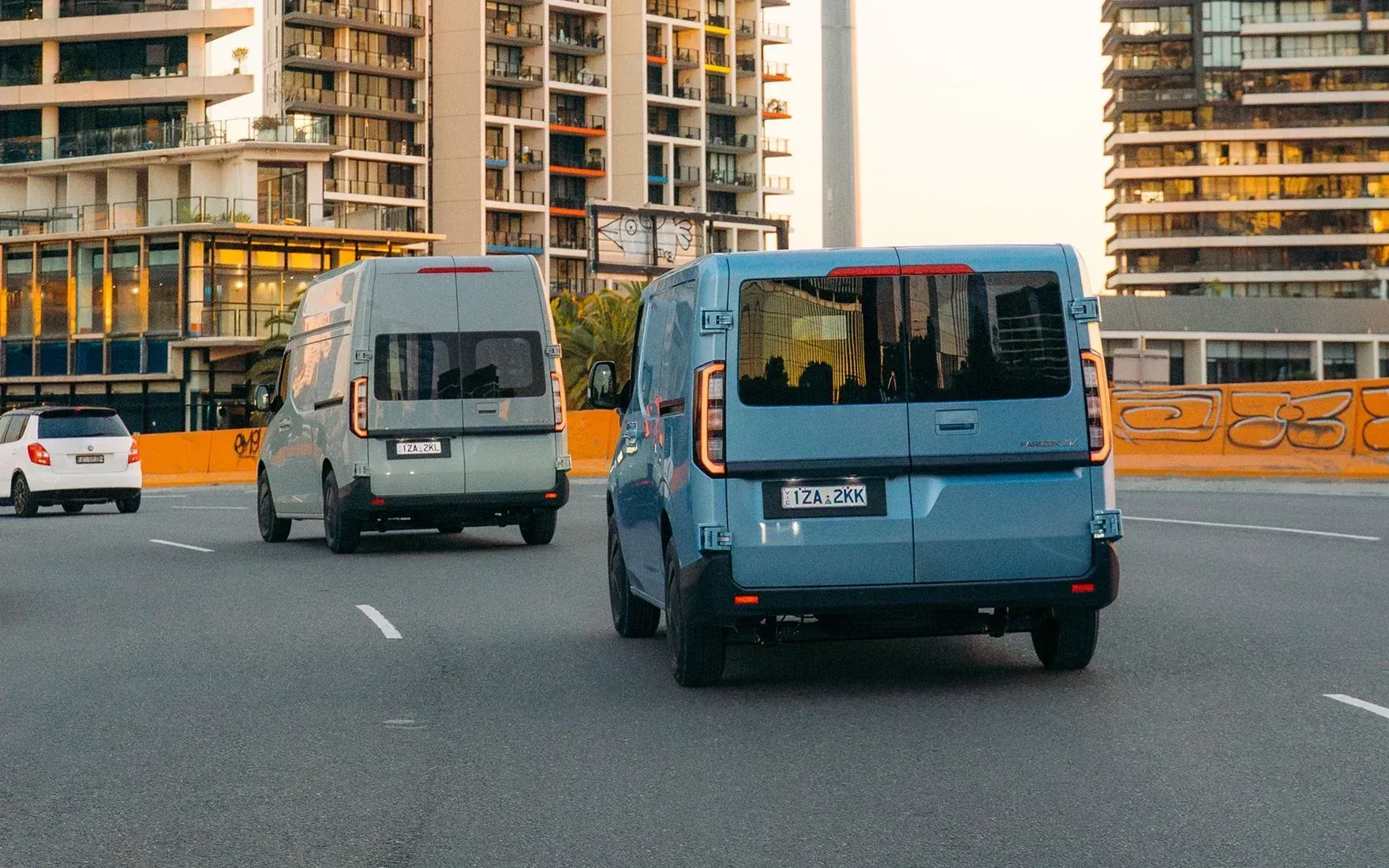Two vans, one light gray and one blue, drive on a city road lined with tall apartment buildings.