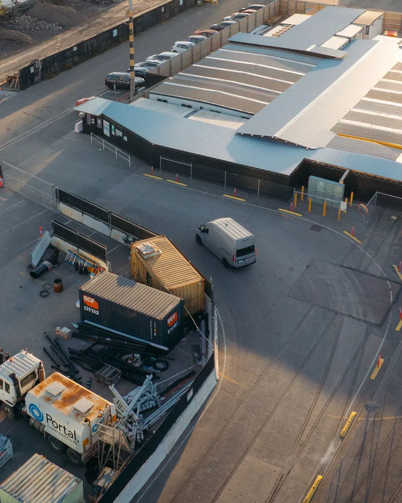 An aerial view of an industrial work site featuring a light-colored van driving past storage containers and a facility.