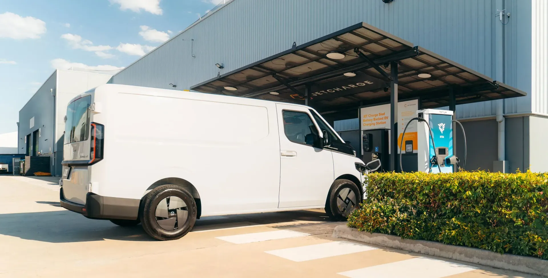 A white electric delivery van is charging at a solar-covered station next to a large warehouse building.