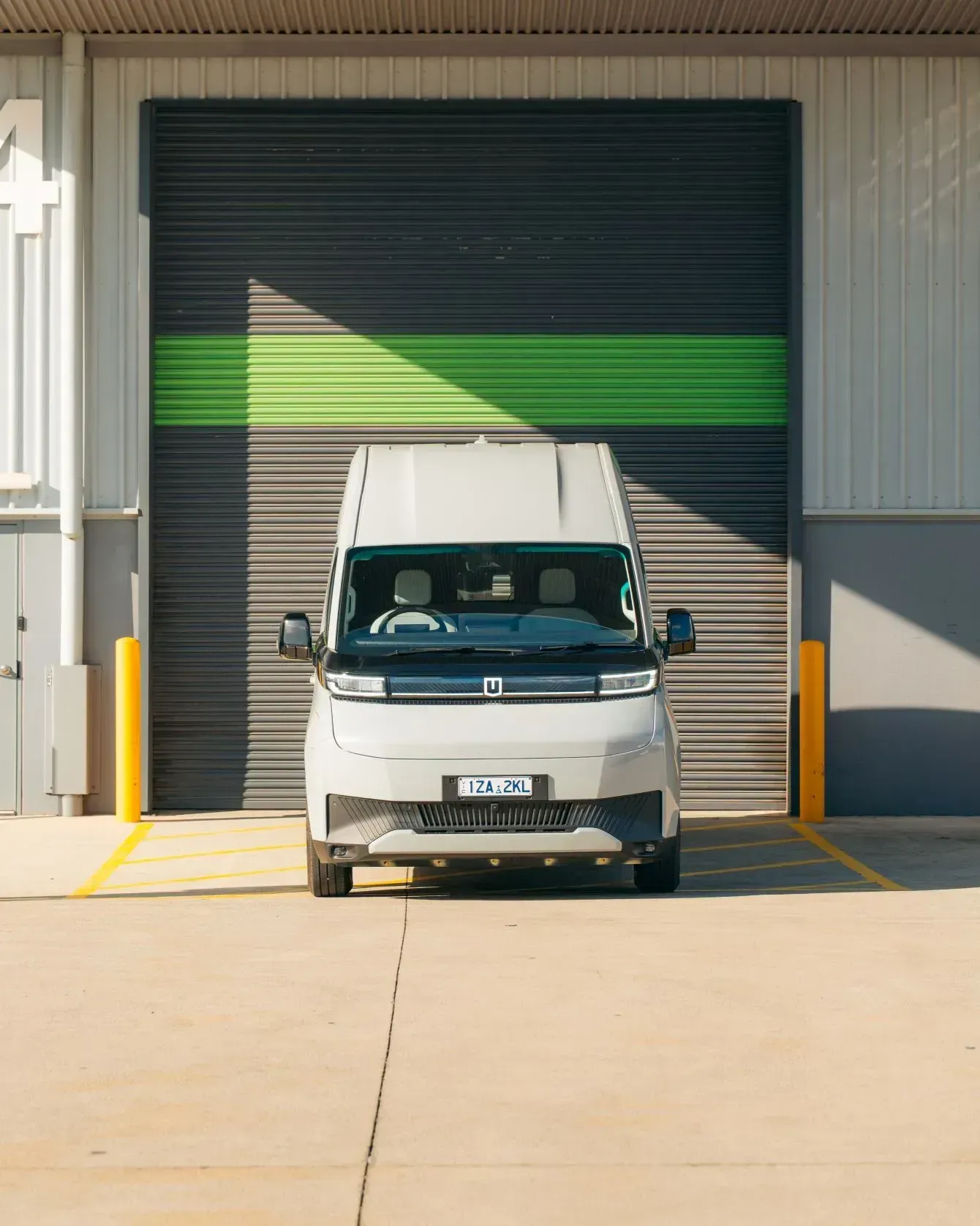 A light gray commercial electric van parked in front of a warehouse door with horizontal black and green stripes.