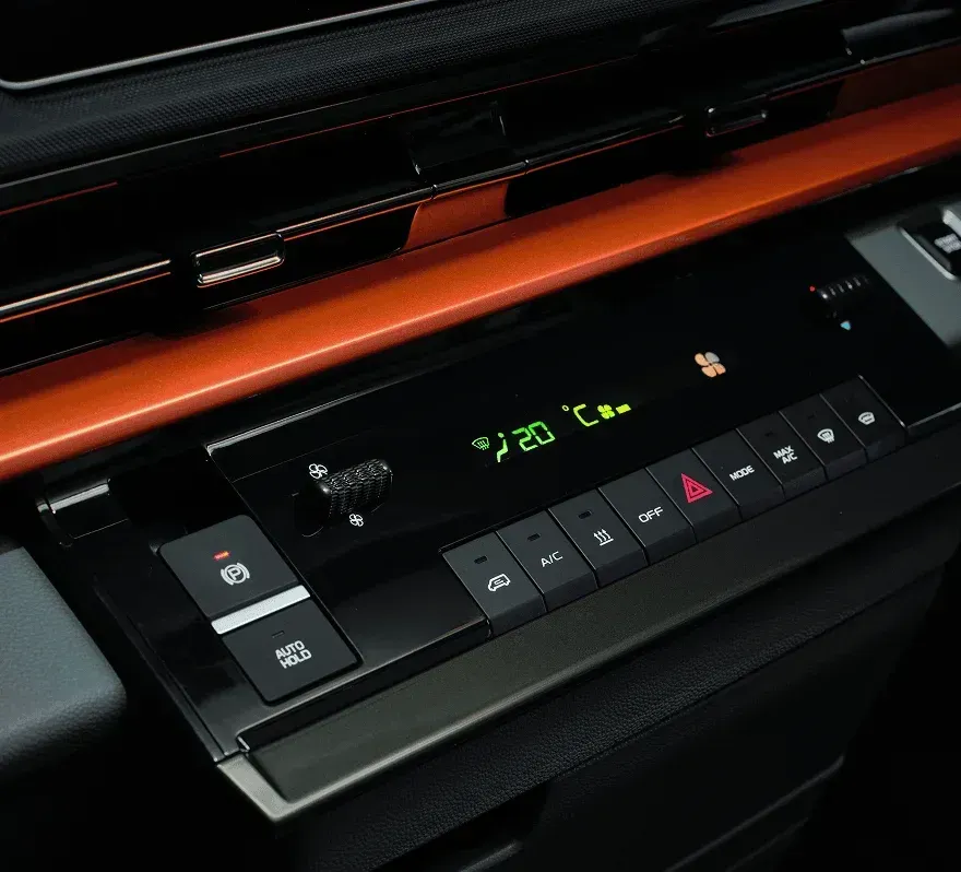 A black car interior console with a green digital climate control display, touch buttons, and an orange trim piece.