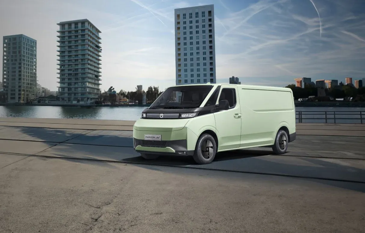 A white electric cargo van parked on a paved lot beside a long, industrial corrugated metal wall under bright sunlight.