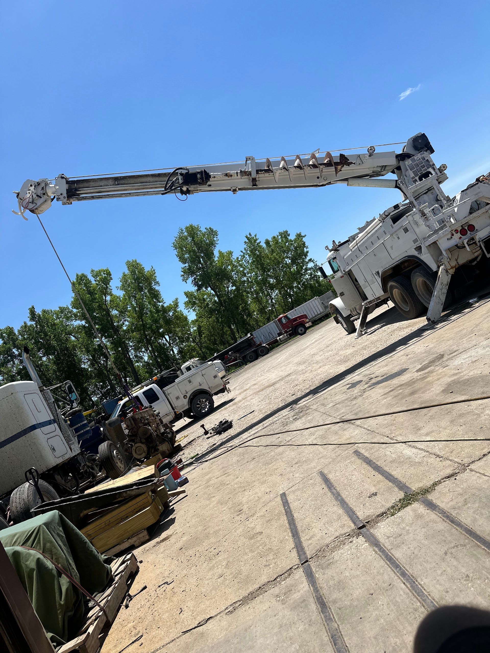 A white crane truck extends its arm against a blue sky, parked on a gravel lot near trees | Doyle Diesel