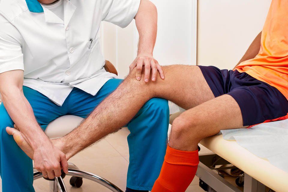 Doctor examining a soccer player's knee in a clinic. Player wears orange shirt, doctor blue pants/white coat.