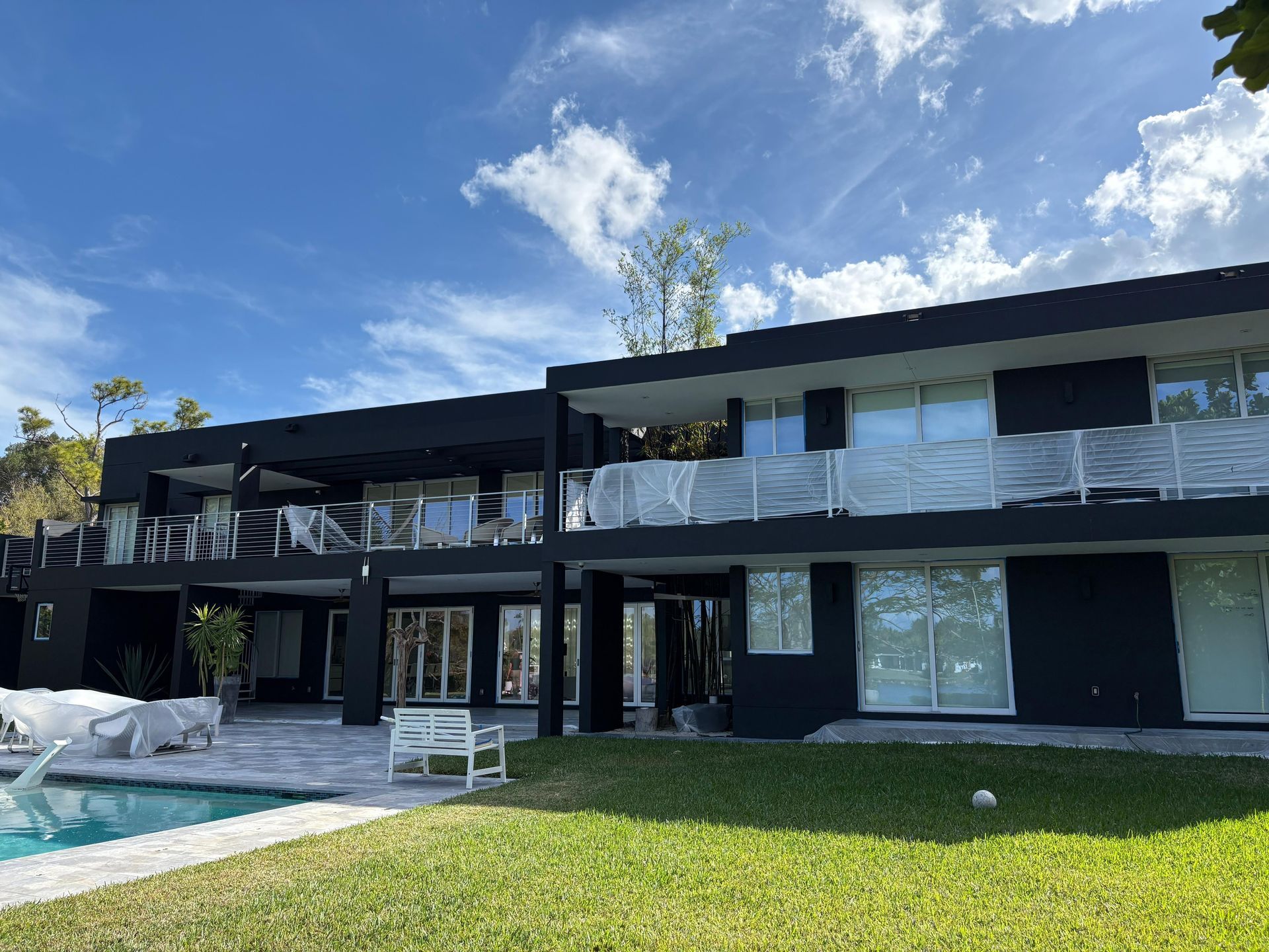 Black modern home with pool, balconies, and lawn against a blue sky with clouds.