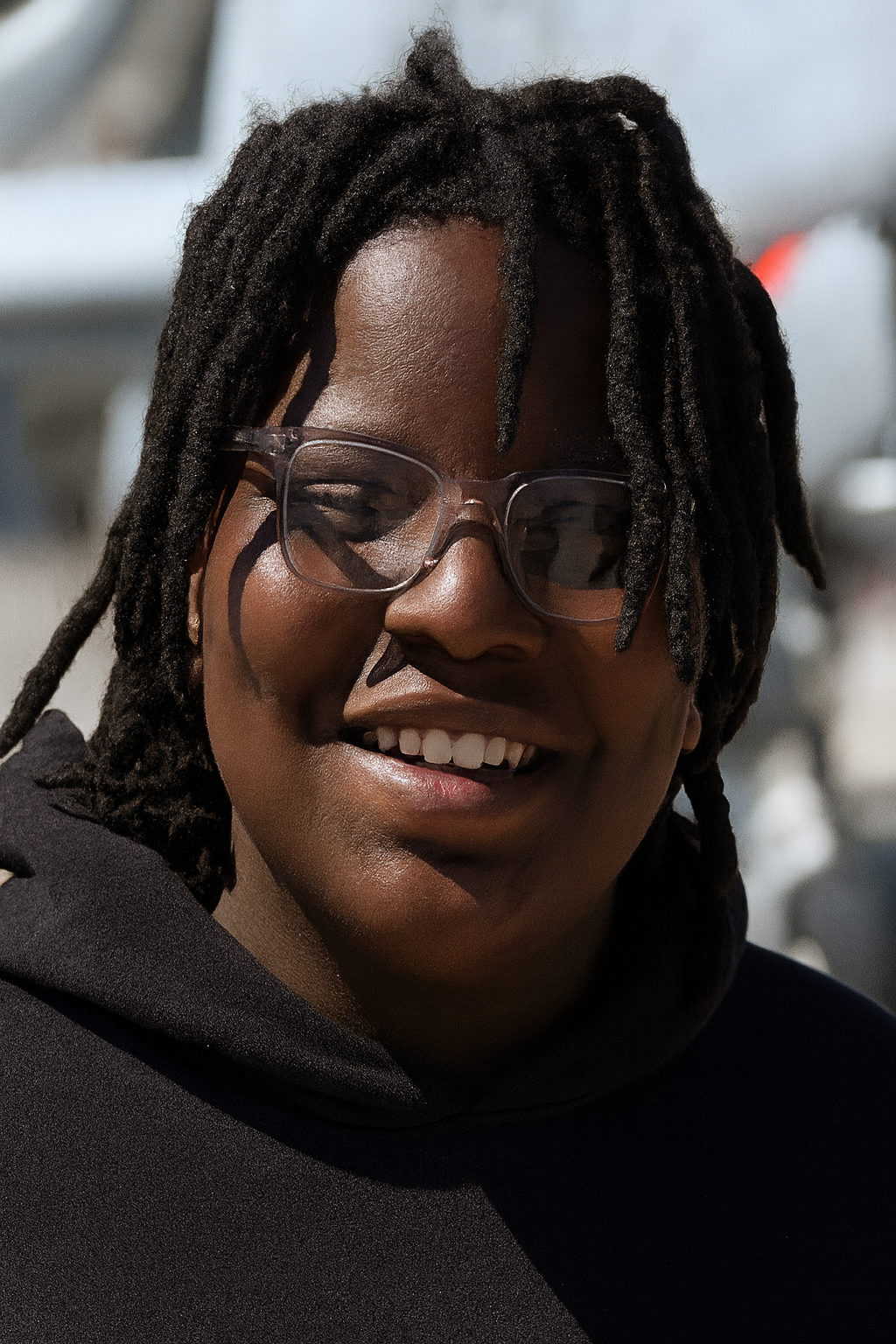 Person with dreadlocks and glasses smiles, outdoors.