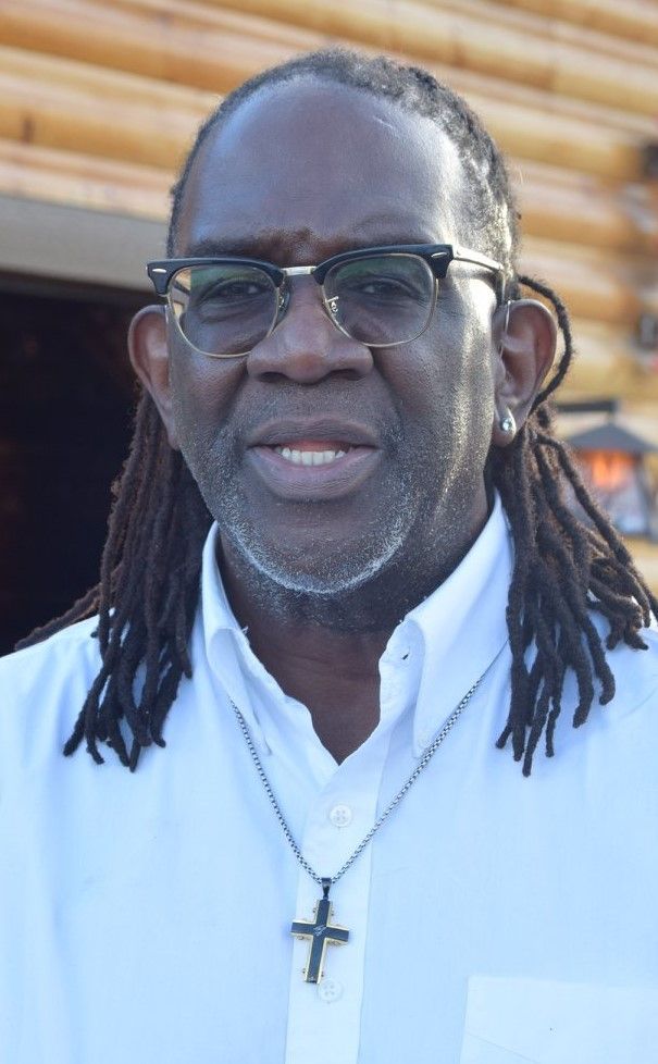 A man with dreadlocks and glasses is wearing a white shirt and a cross necklace.