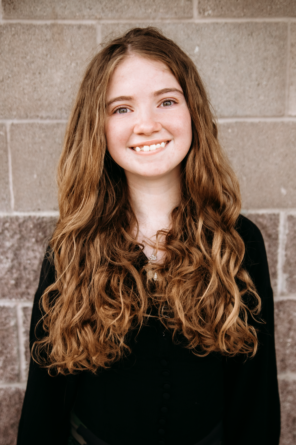 Young woman with long, wavy auburn hair smiling, standing in front of a brick wall.
