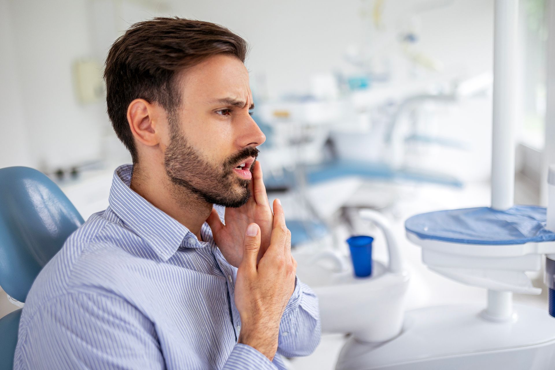 Man in dentist chair, grimacing and holding his jaw. Light-blue chair, dental equipment in background.