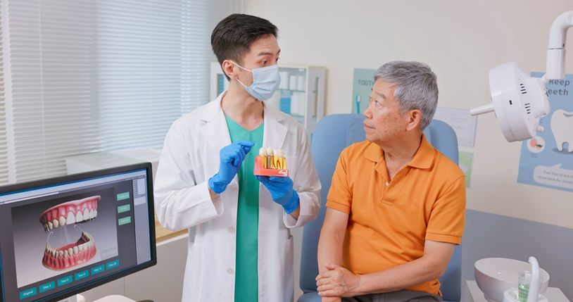 Dentist showing dental X-ray to seated patient in clinic, with monitor displaying teeth model