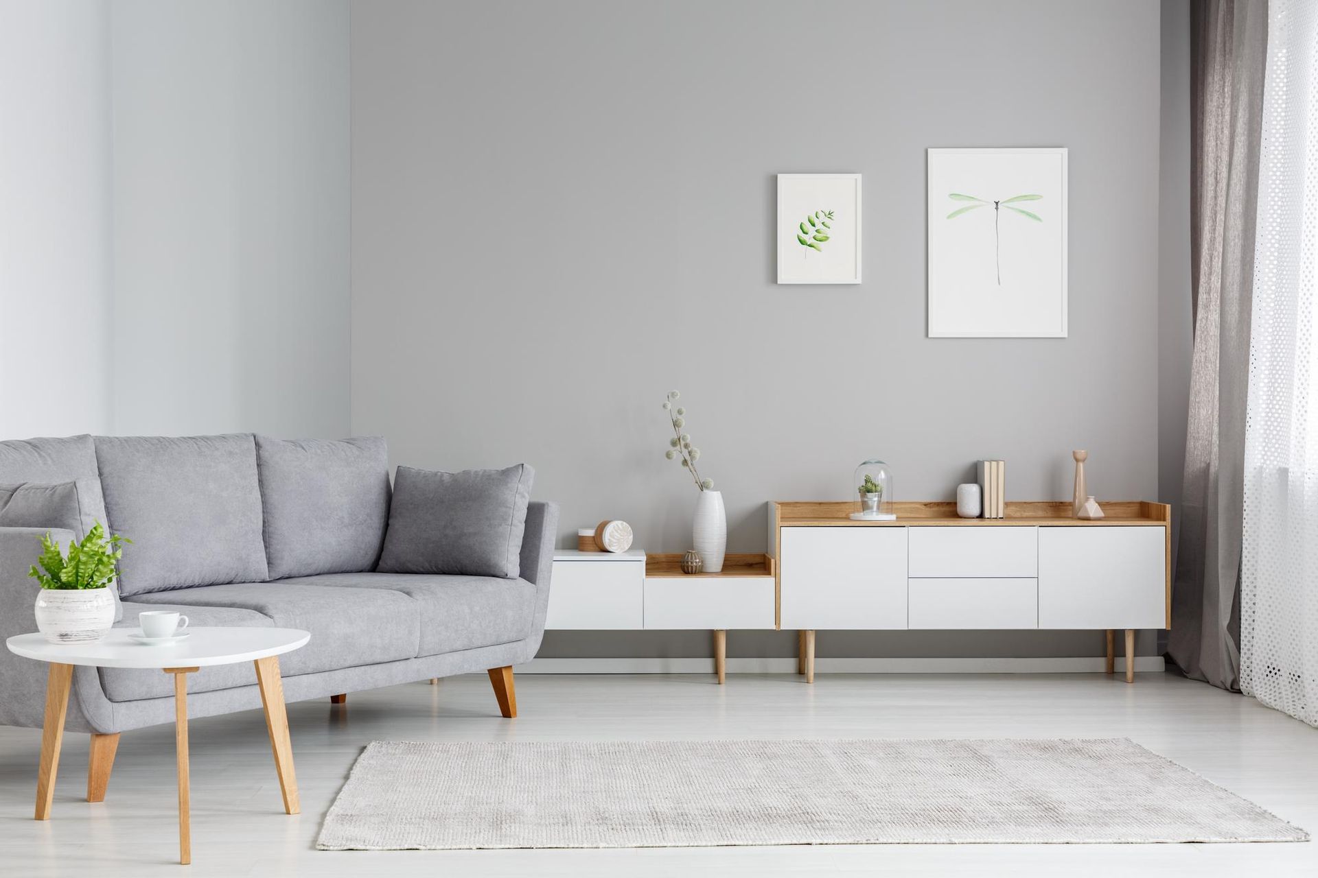 Gray living room with a couch, table, rug, and cabinet against a gray wall with two framed prints and a sheer curtain.