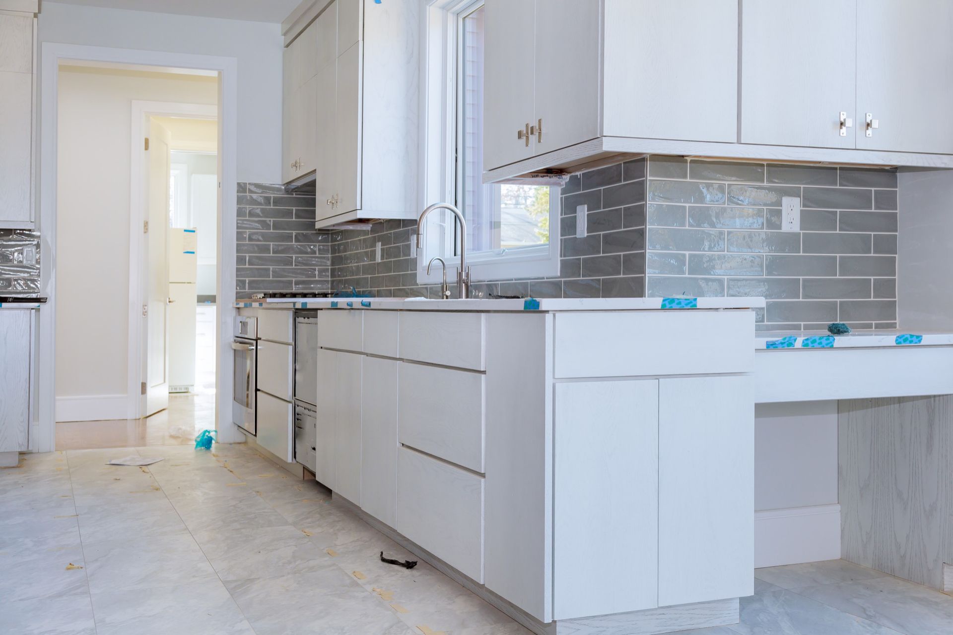 White kitchen cabinets and counters, gray backsplash, sink, and window.