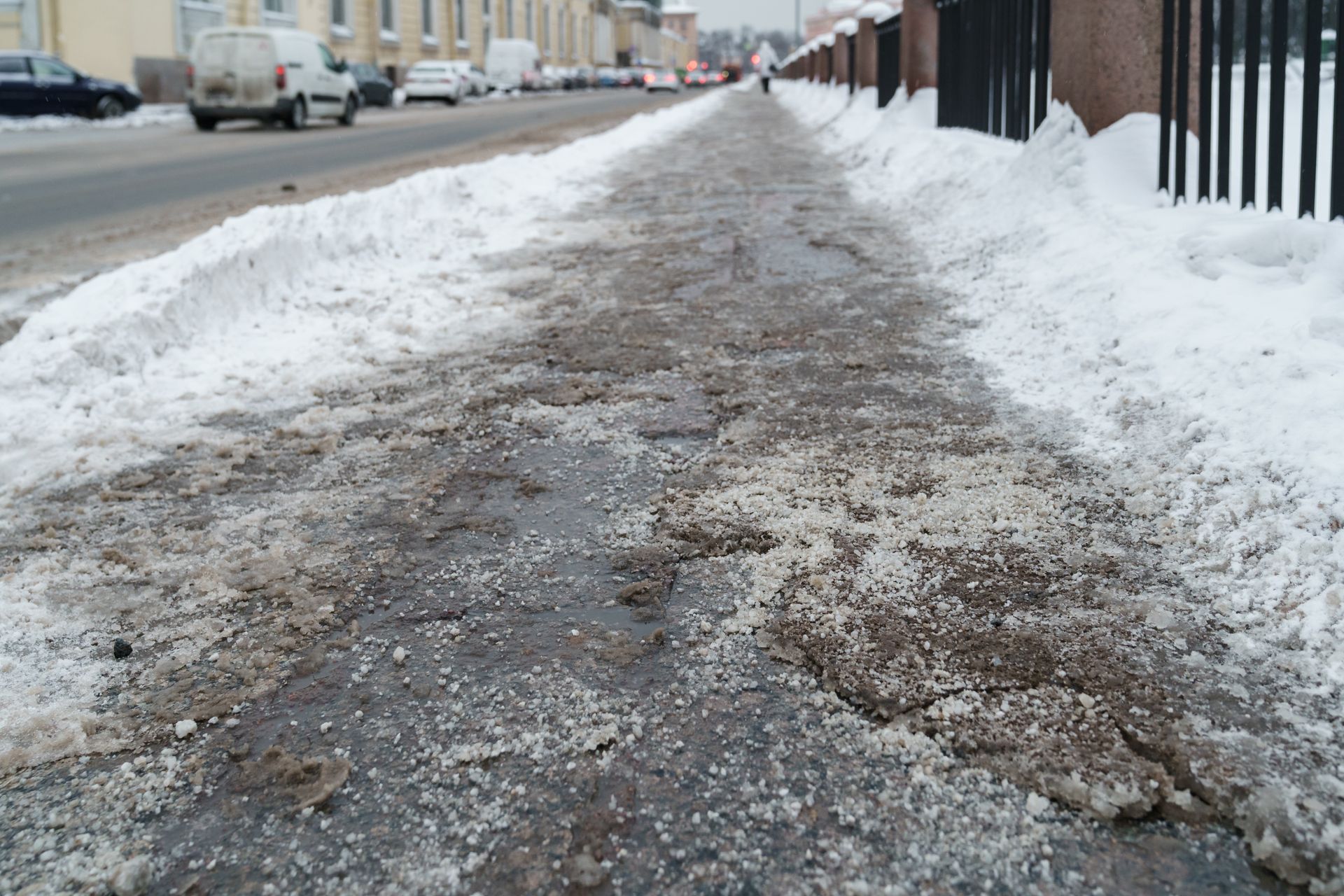 Snowy concrete sidewalk with rock salt, slush, a street, and a fence.