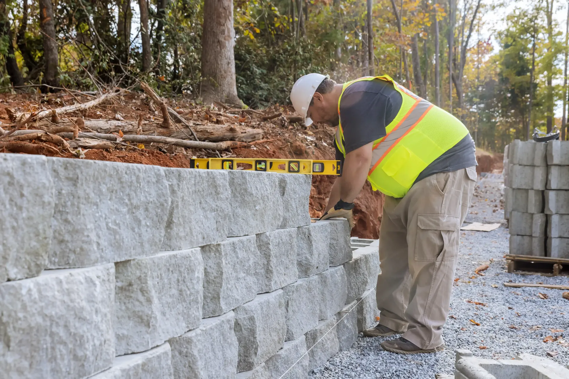 Construction worker in safety vest using a level on a retaining wall made of concrete blocks outdoors.