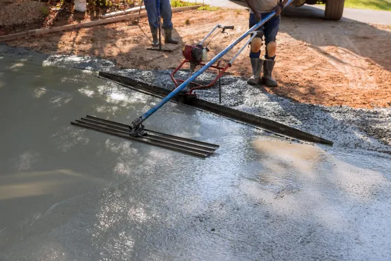 Person using a concrete screed to level wet concrete. Outdoor setting with tools and gravel.