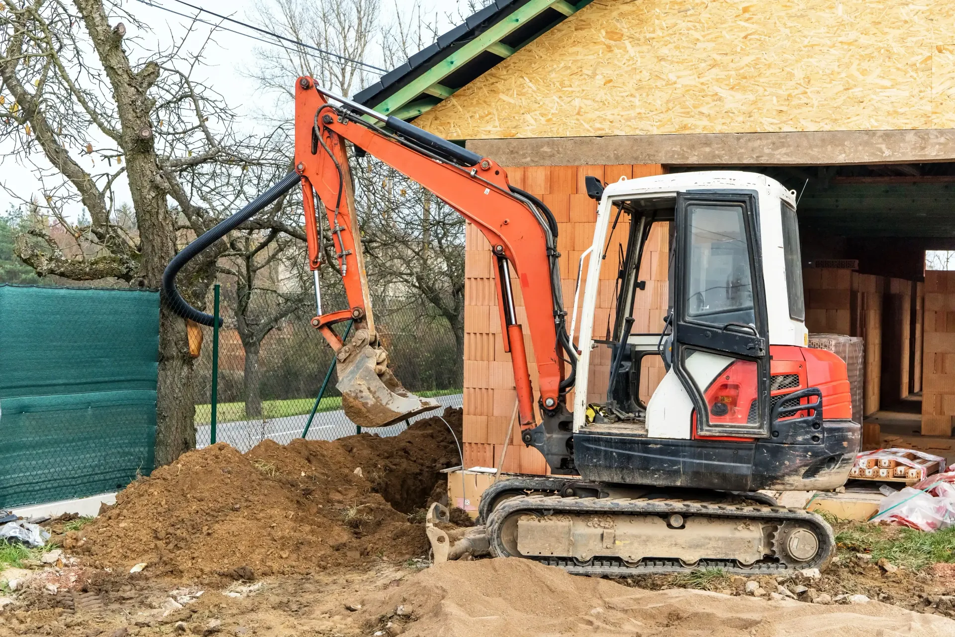 Orange and white excavator digging a trench next to a house under construction.