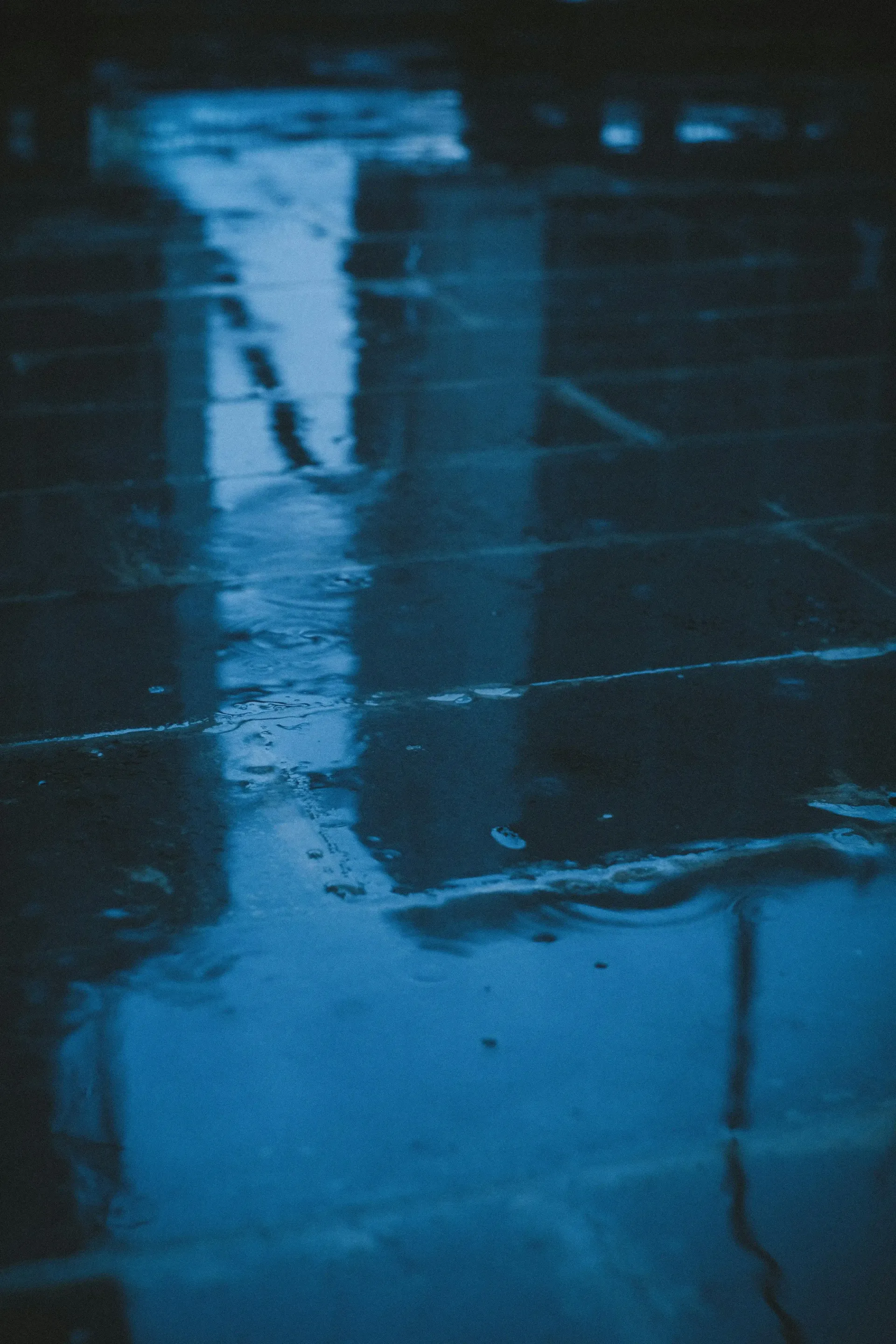 Puddle on concrete reflects blurry architecture, illuminated in blue.