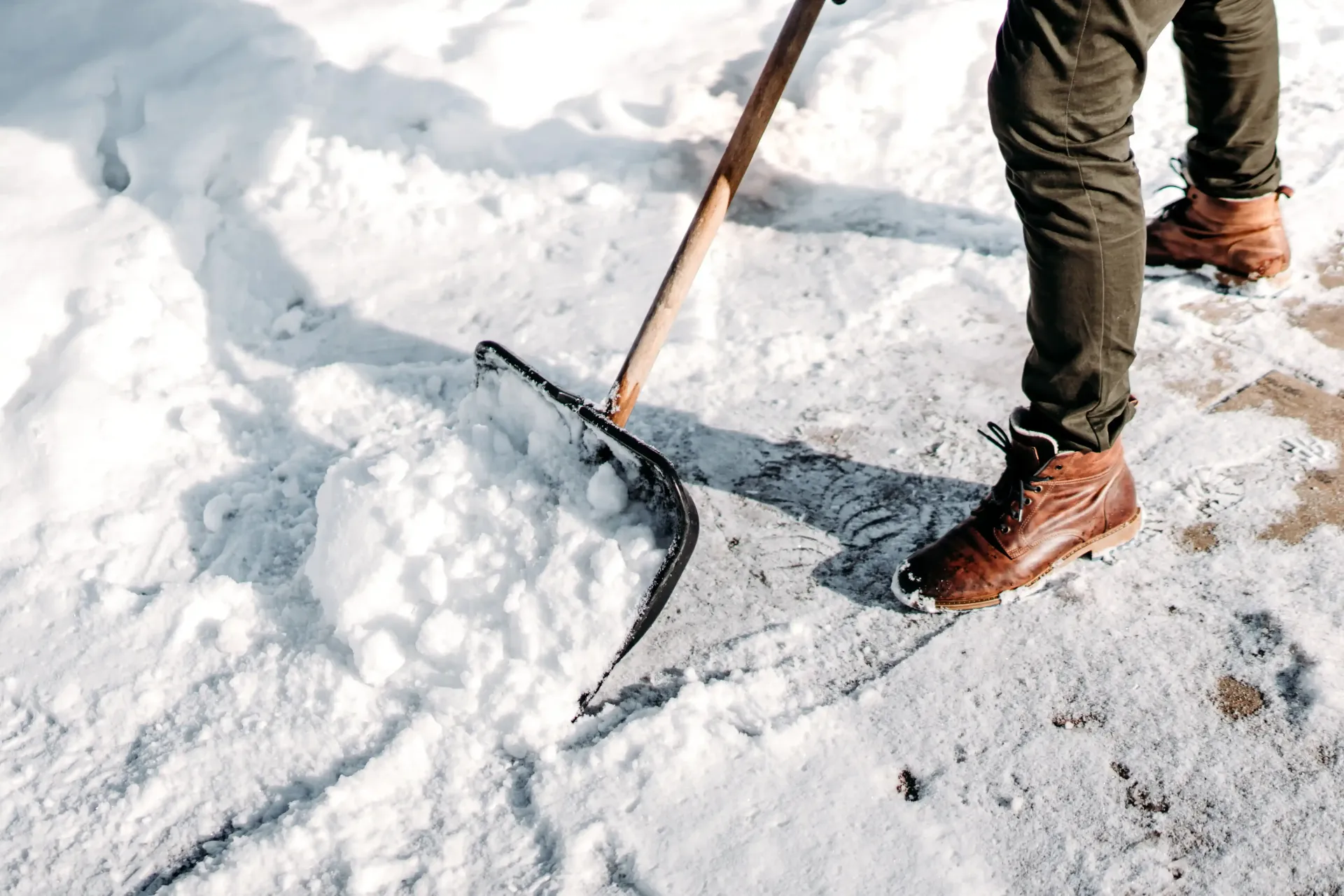 Person shoveling snow on a concrete surface, wearing brown boots and dark pants; snow is white.