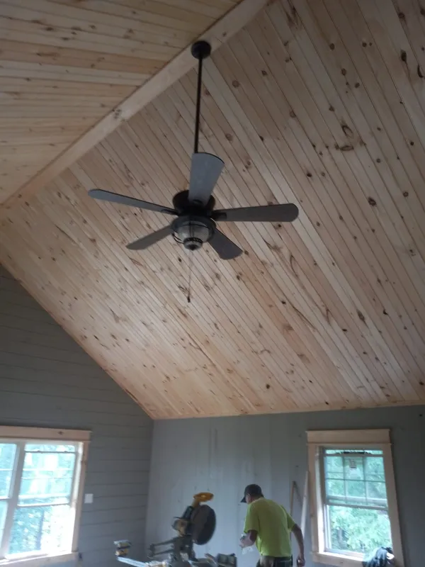 Room with vaulted wood ceiling, ceiling fan, and two windows. Man working at a saw is in the lower center.