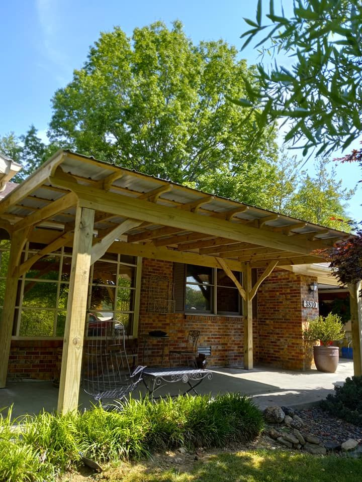 Wooden pergola over a brick patio, with windows, a chair, and greenery under a sunny sky.