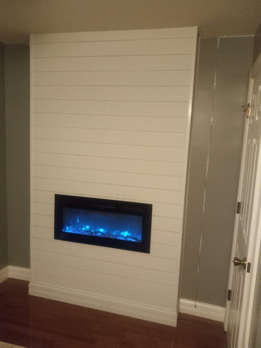 White shiplap fireplace surround with electric fireplace and blue flames. Brown floor, gray walls, white door.