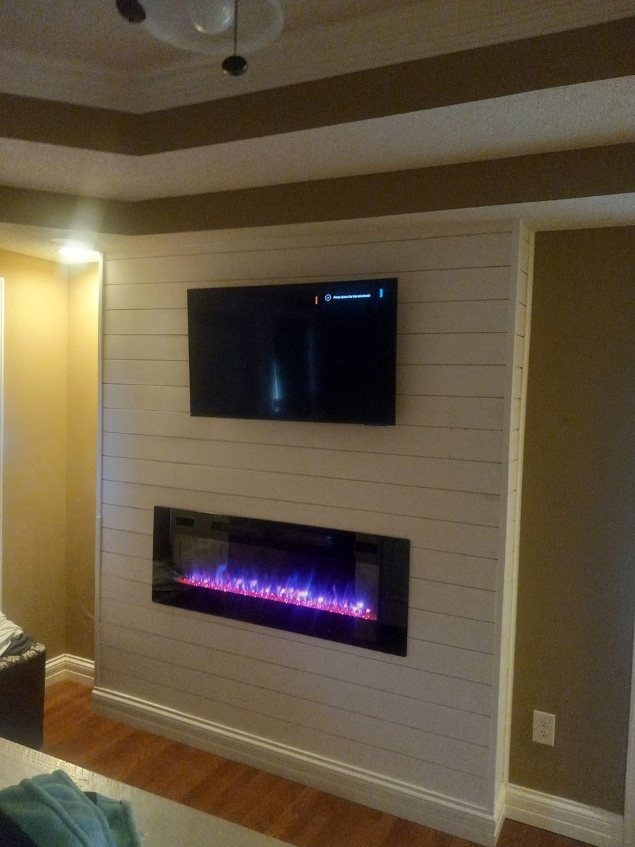 TV mounted above an electric fireplace on a white shiplap wall, in a room with brown accents.