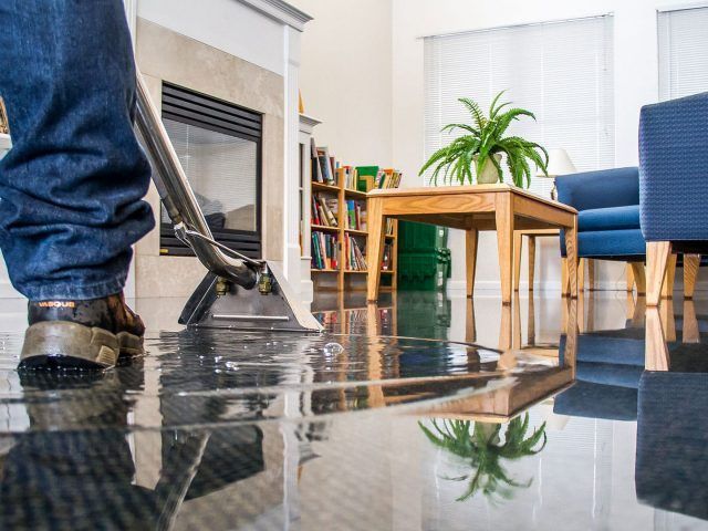 Person cleaning a wet floor with a machine; a living room with furniture is in the background.