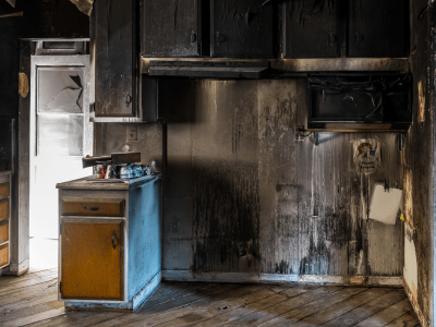Burnt kitchen interior: charred cabinets, walls, and appliances; sunlight streams in doorway.