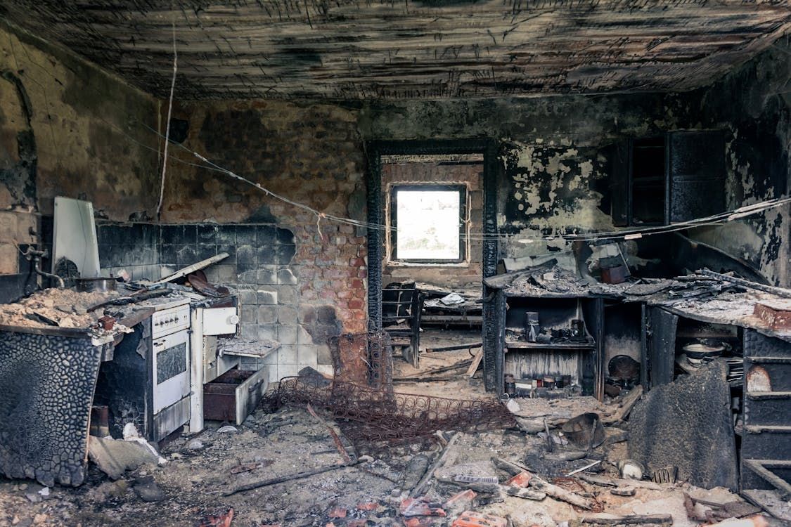 A kitchen destroyed by fire: charred walls, appliances, and doorway, with a window at the end of a hallway.
