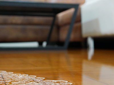 Close-up of glossy wooden floor with water pooling. A dark-framed table and brown sofa are in the background.