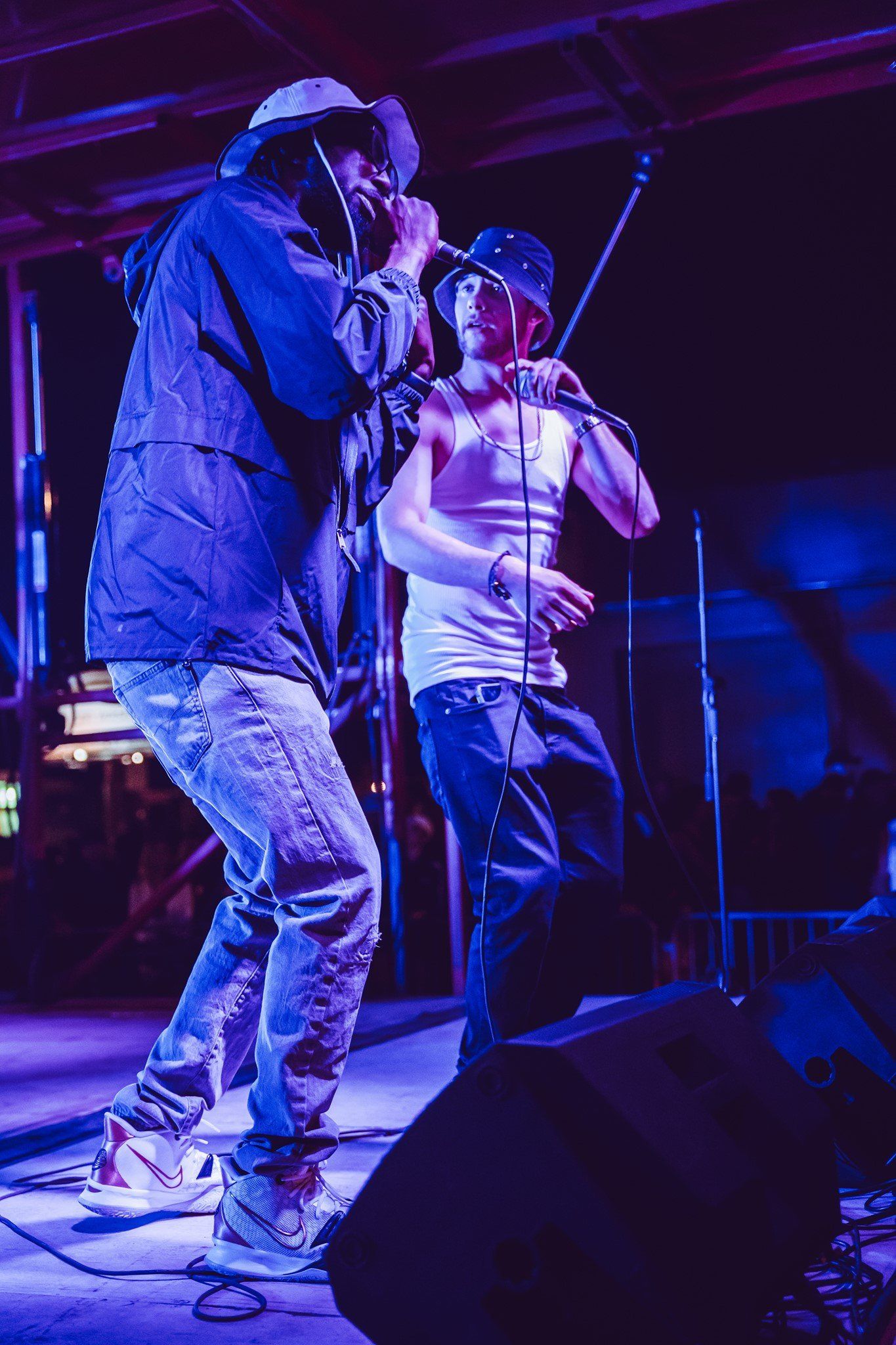 Two men performing on stage under purple light. One in dreadlocks, the other in a bucket hat.