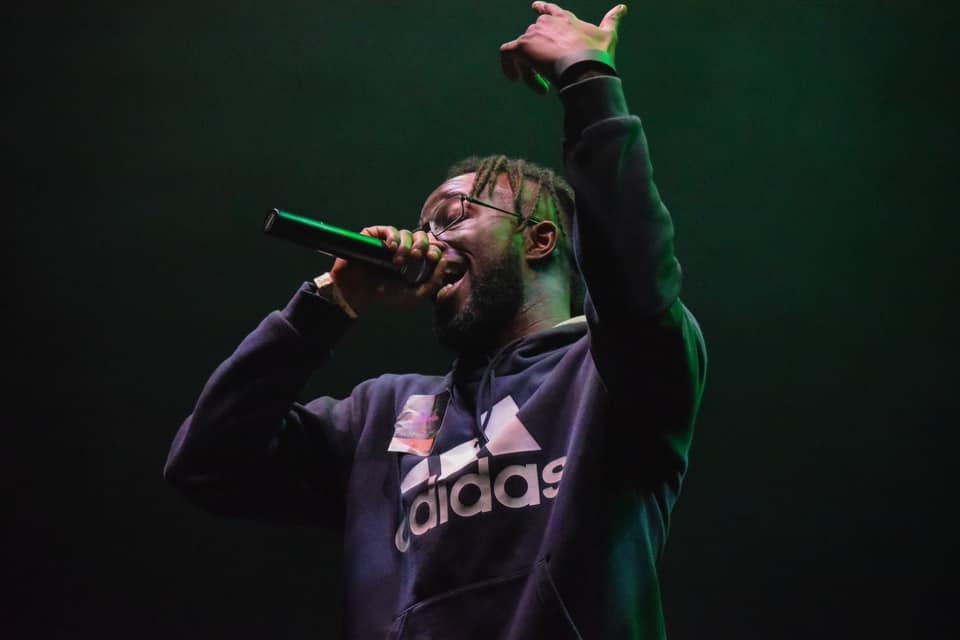 A Black man with glasses, Adidas hoodie, sings into a mic on stage, green lighting.