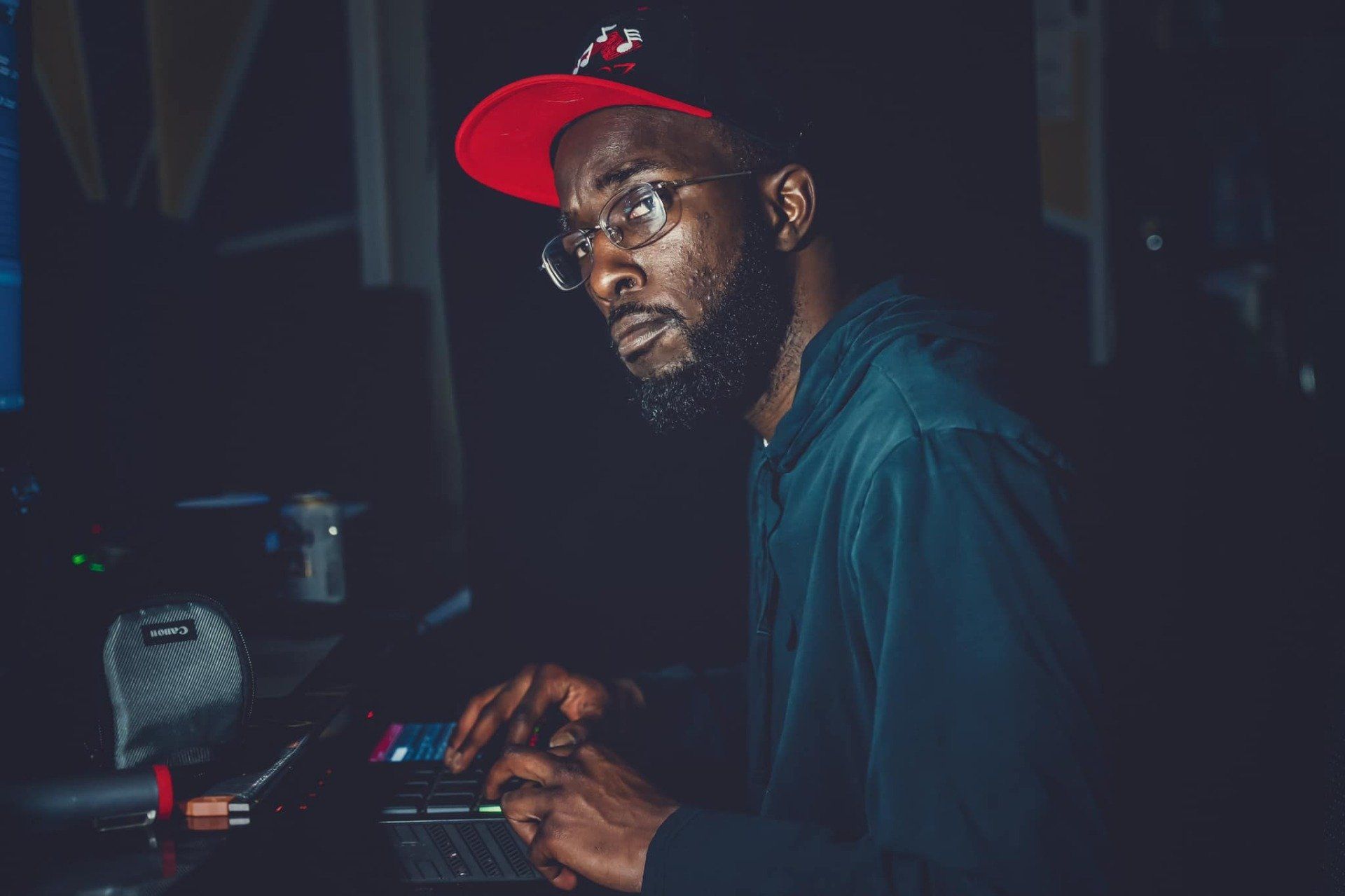 Man in red cap and glasses working on a laptop, looking to the side. Dark setting.