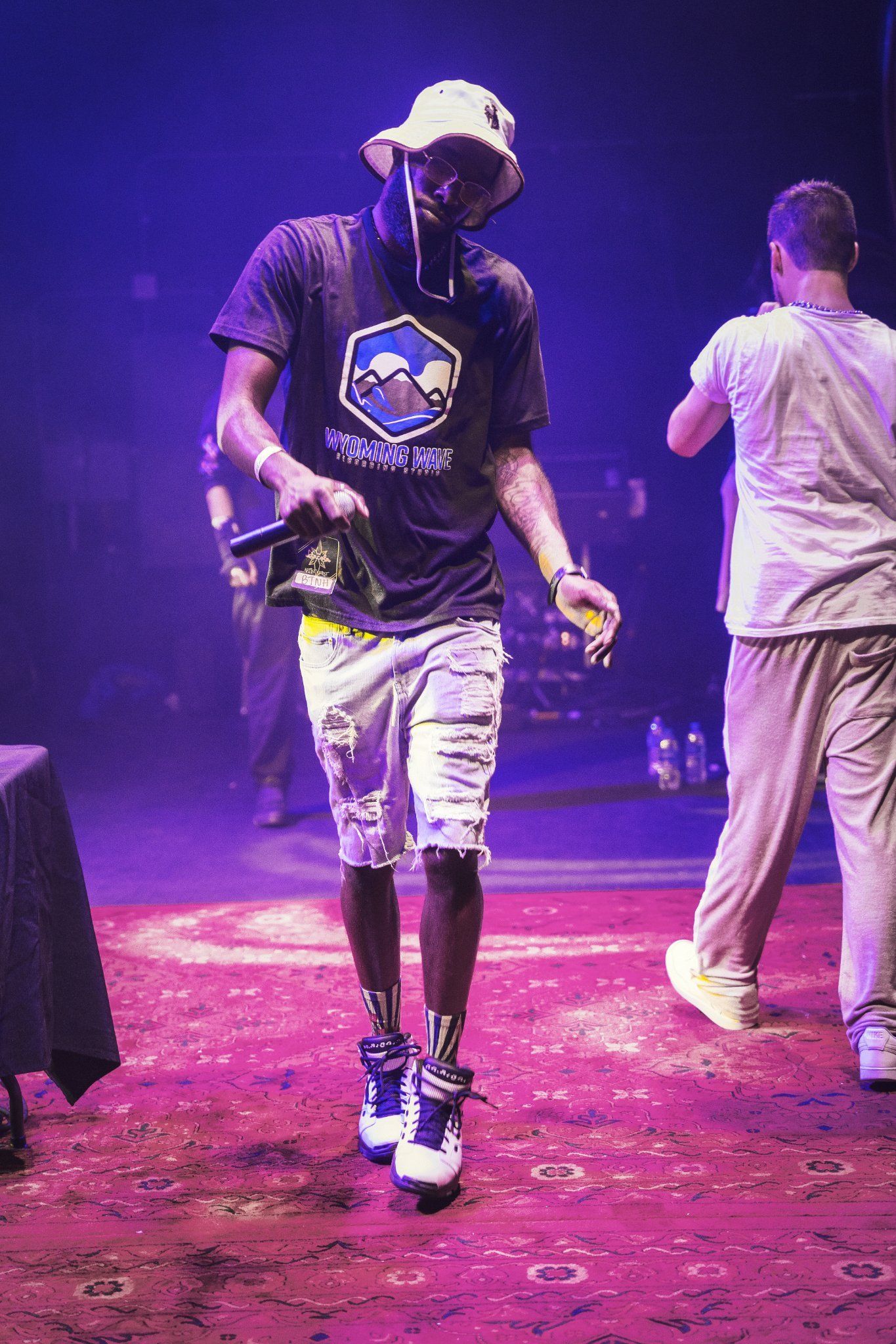 Man on stage wearing hat, shirt, and shorts, holding a microphone. Another man in background.