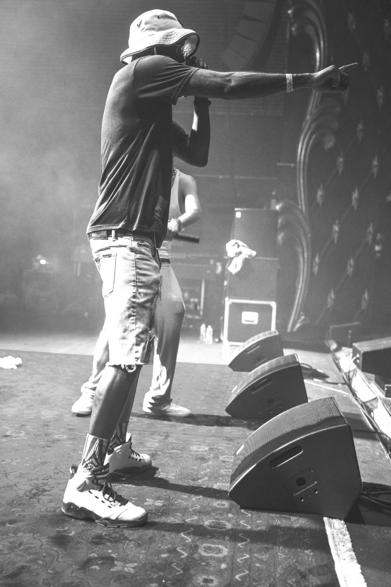 Rapper on stage, pointing with mic. Wearing bucket hat, t-shirt, shorts, and sneakers, another figure behind.