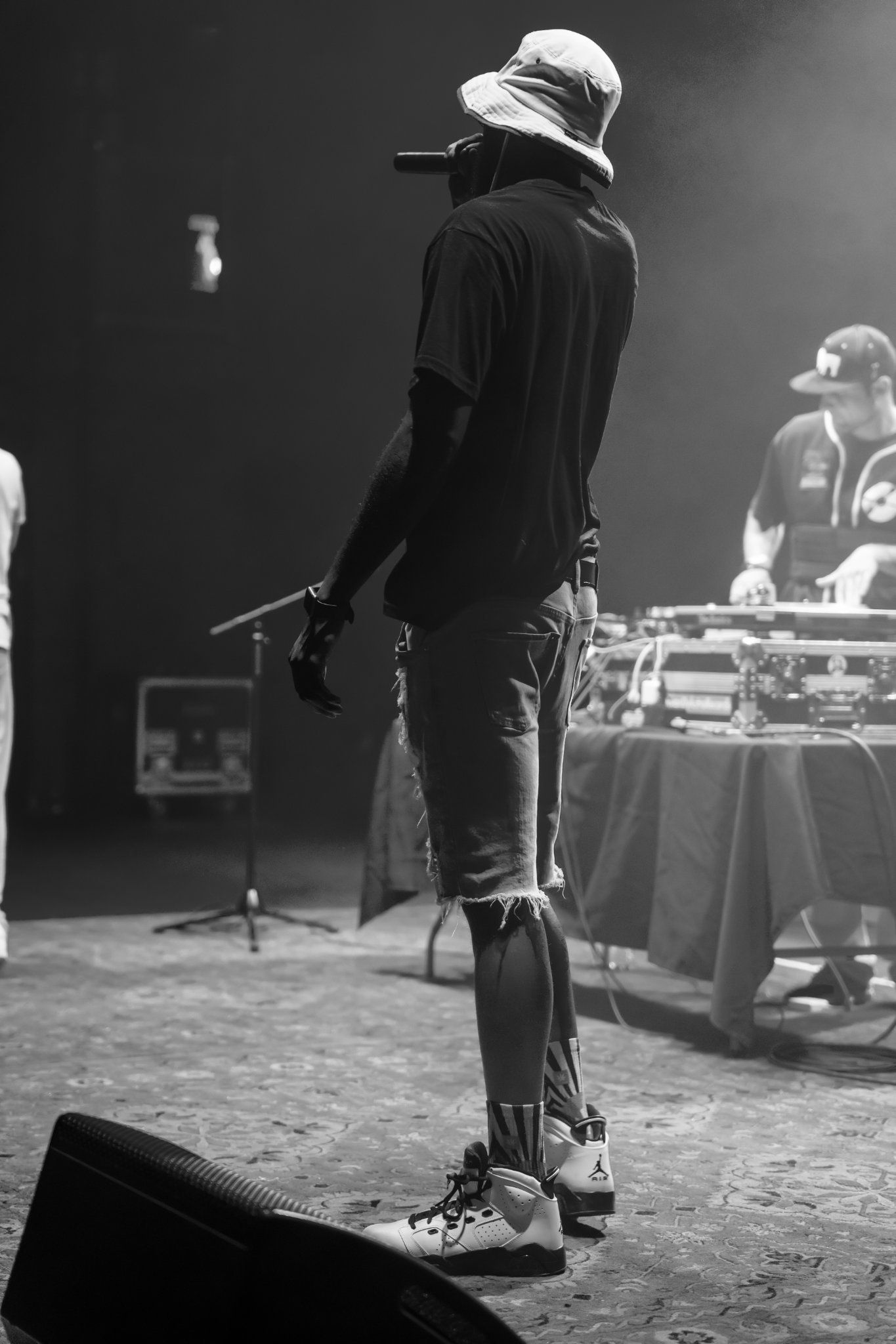 A musician with a bucket hat performs on stage with DJ equipment and another person in the background.