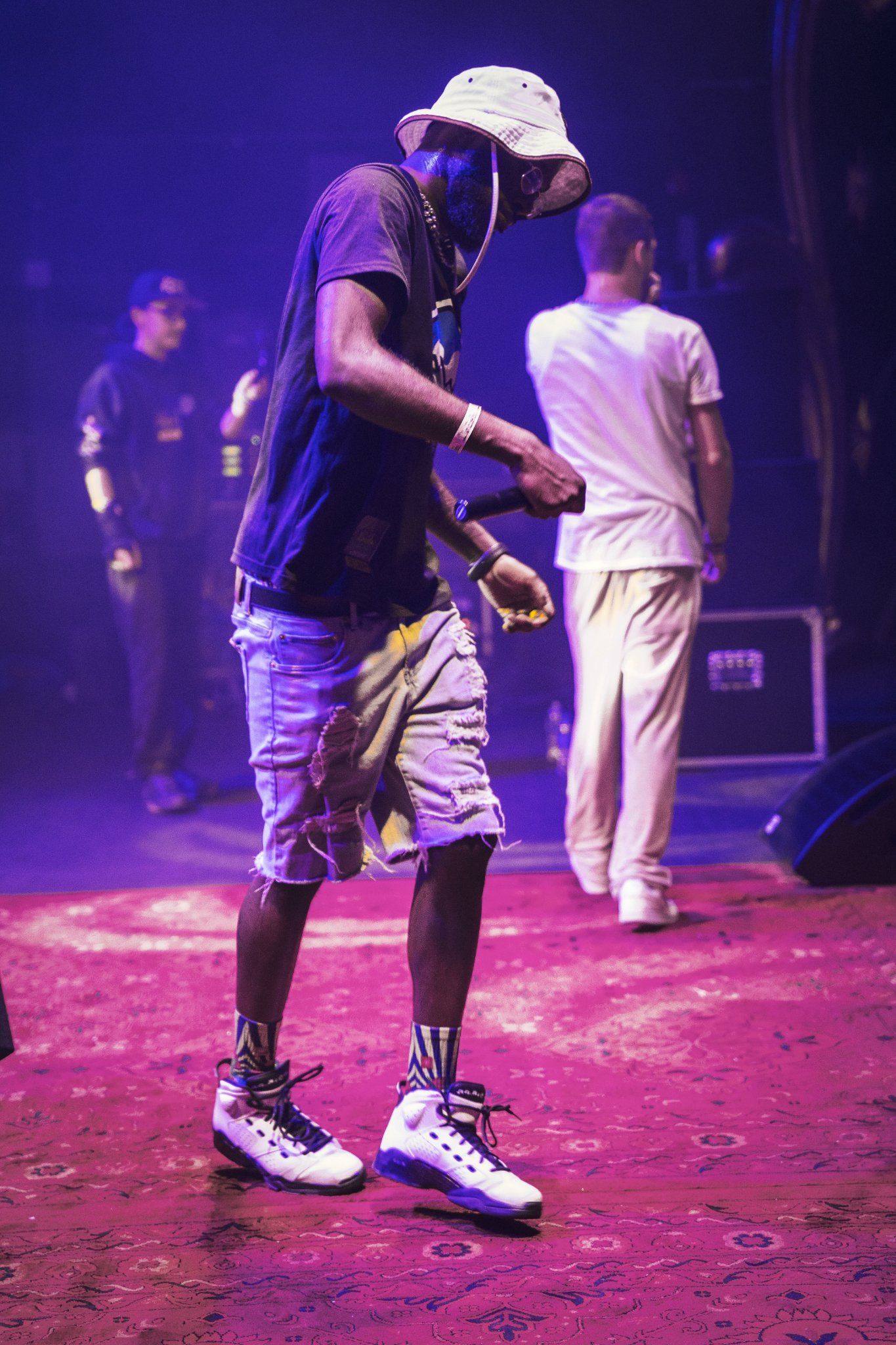 A person with hat, shorts, and sneakers performs on stage under blue light.