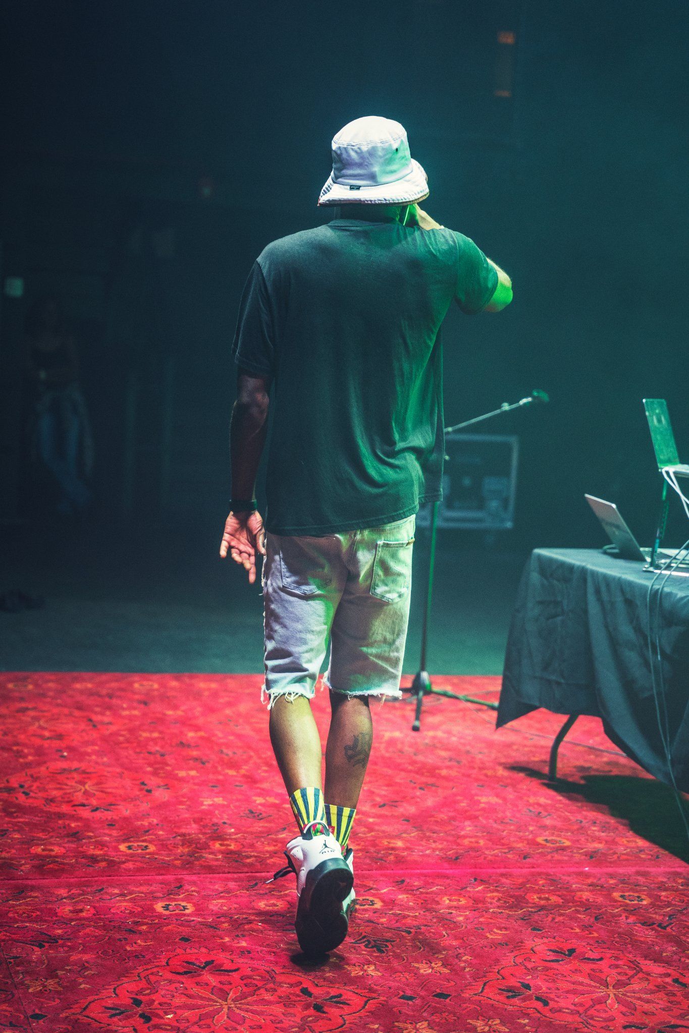 Man in a hat walking away on a red carpeted stage, dark background, microphone and laptop visible.