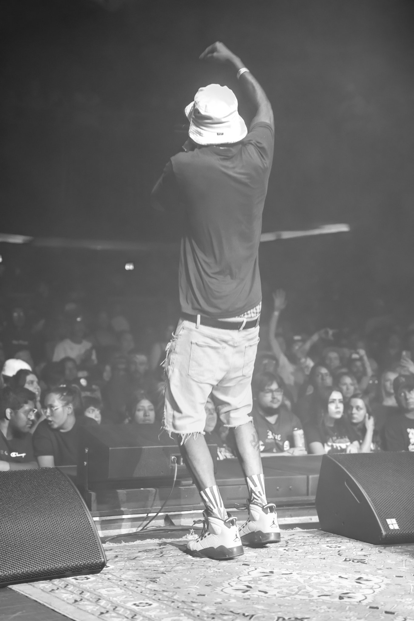 Man in bucket hat on stage performing for a crowd, arms raised, black and white.