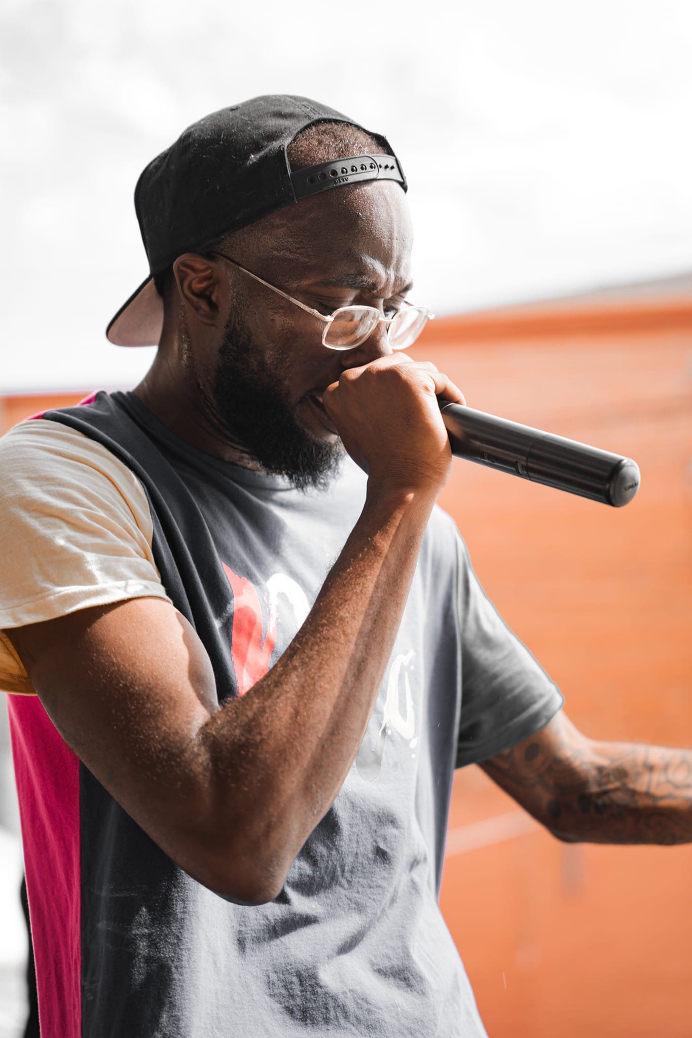 Man with a beard, hat, and glasses, singing into a microphone outside.