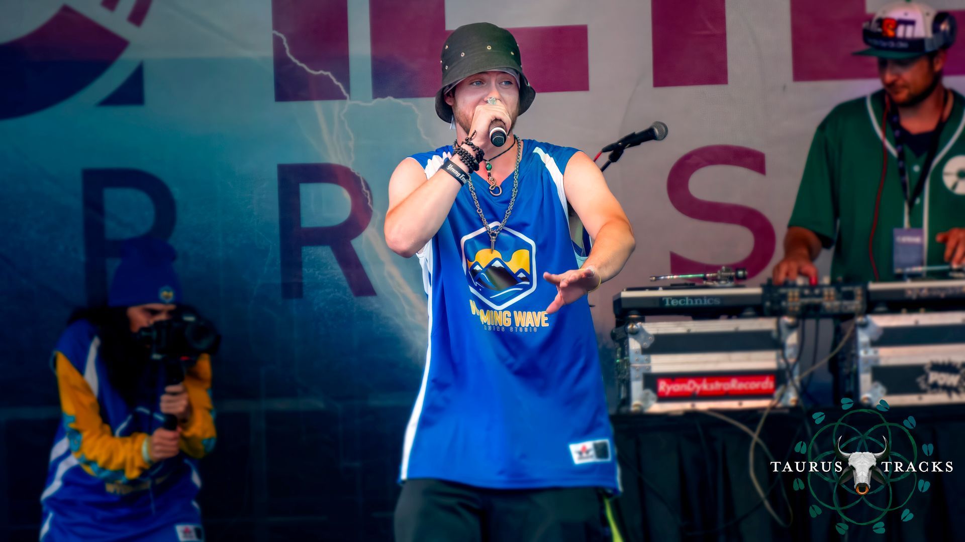 Rapper in a blue jersey performs on stage, gesturing with his hands. DJs work turntables, in a sunny setting.