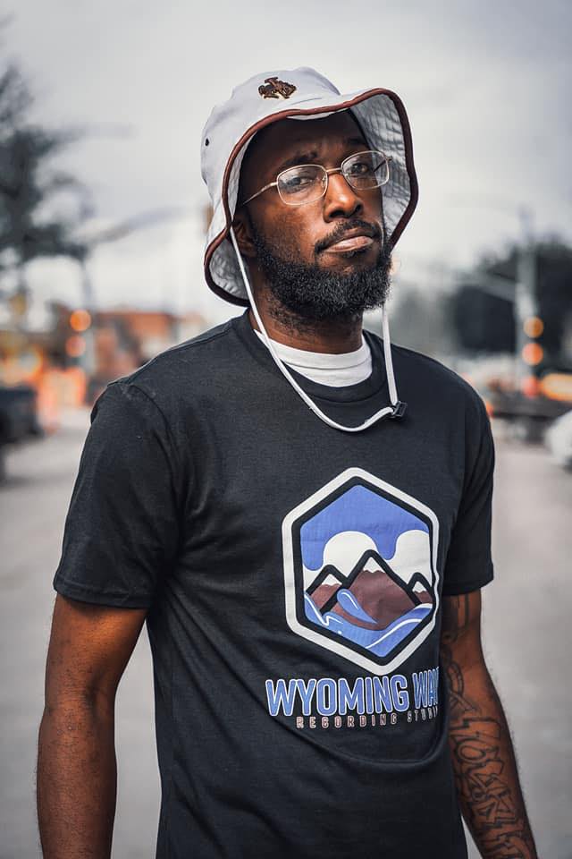 Man with glasses and a bucket hat wearing a shirt with a mountain logo, standing on a street.