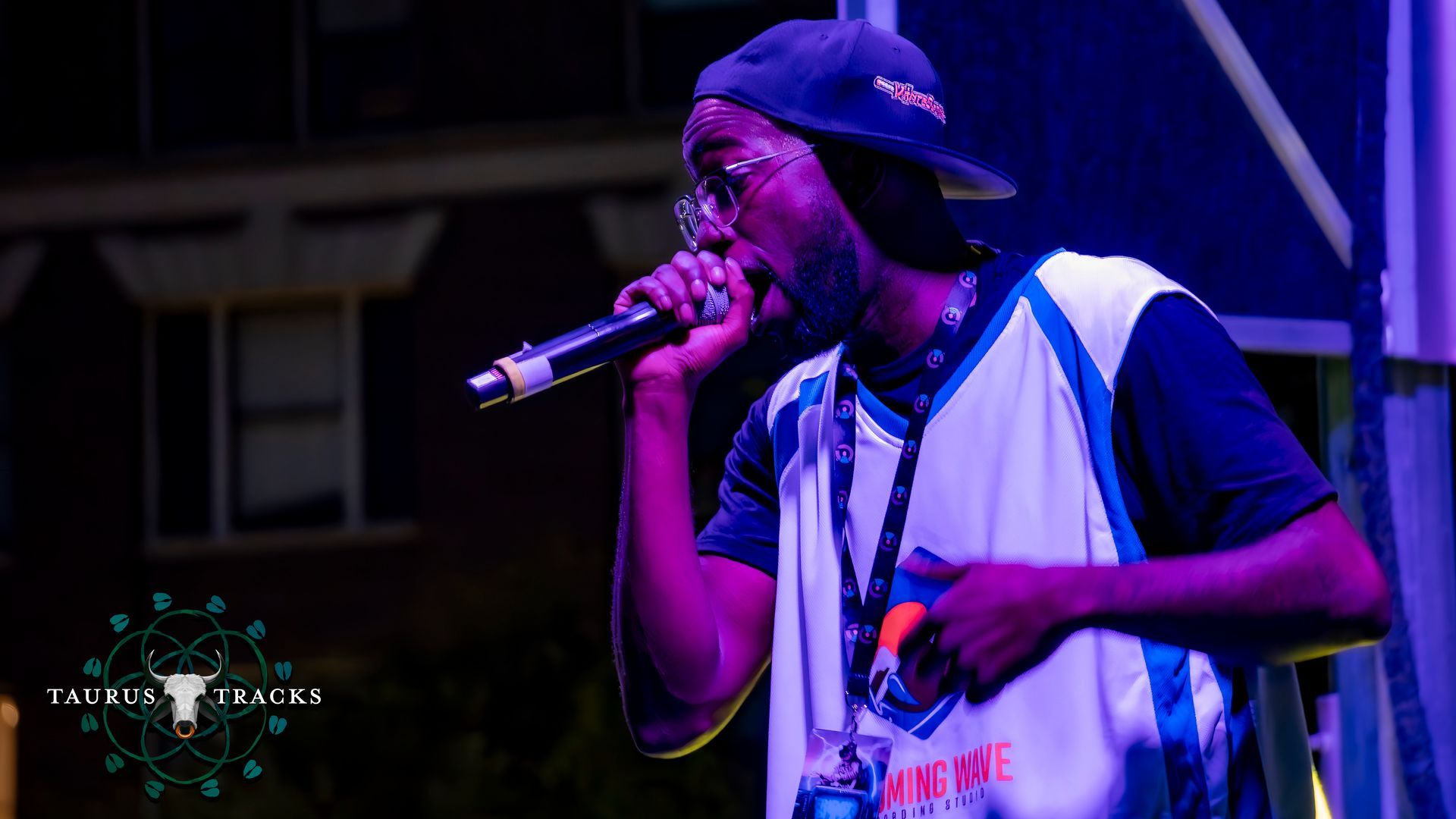 Man in a baseball cap rapping into a microphone on stage, wearing glasses and a basketball jersey. Nighttime performance.