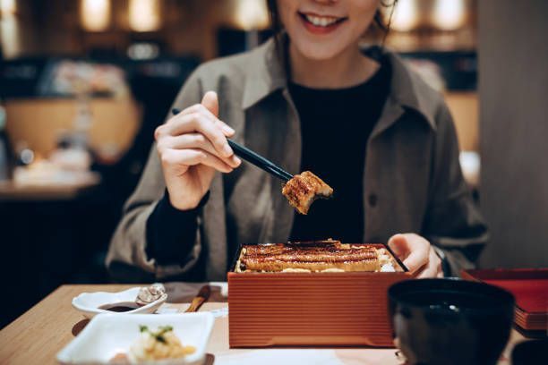 Mid-section of woman enjoying traditional grilled unagi over rice in a bento box in a Japanese restaurant.