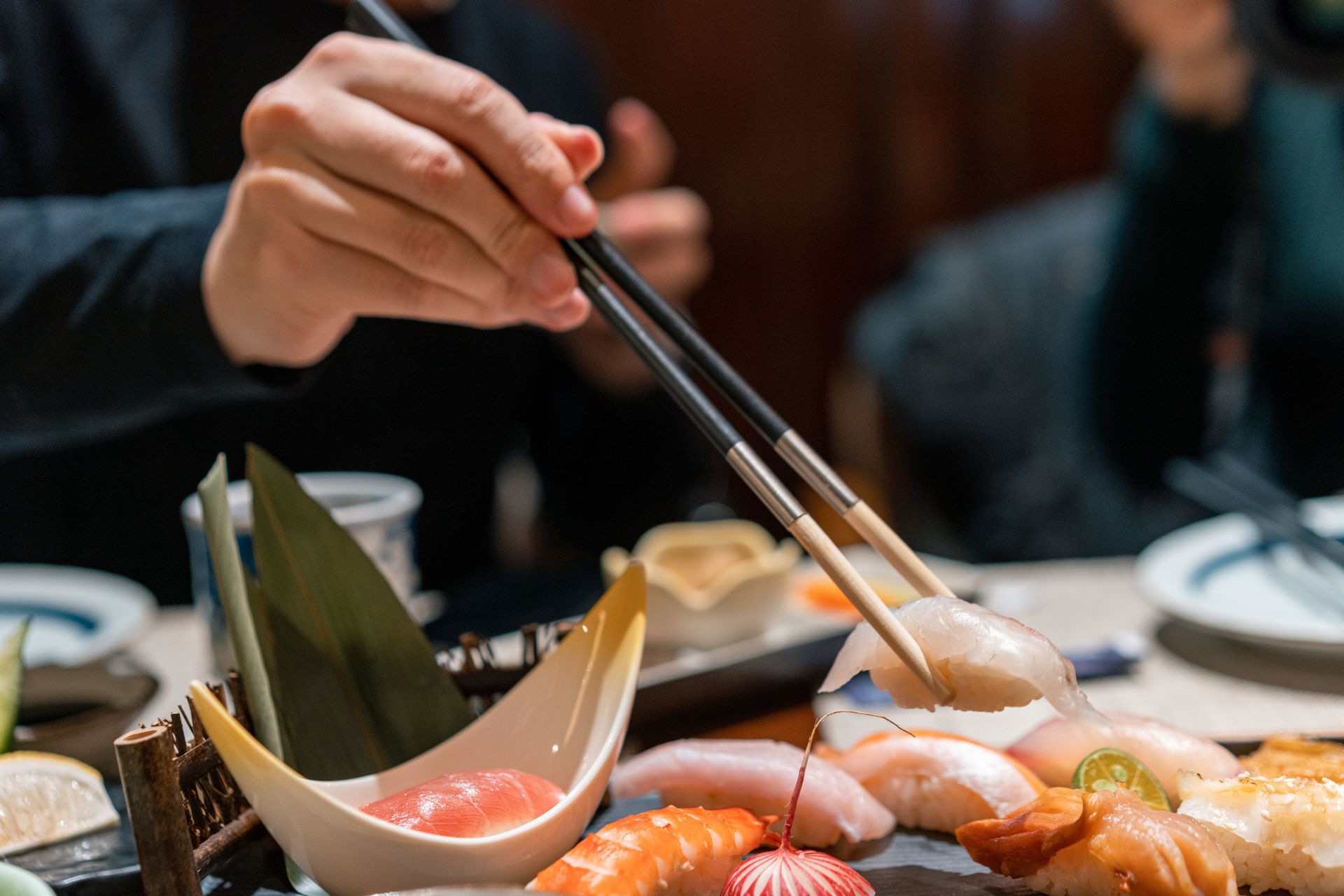 Chopsticks lifting sushi from a platter with nigiri and garnishes in a restaurant setting.