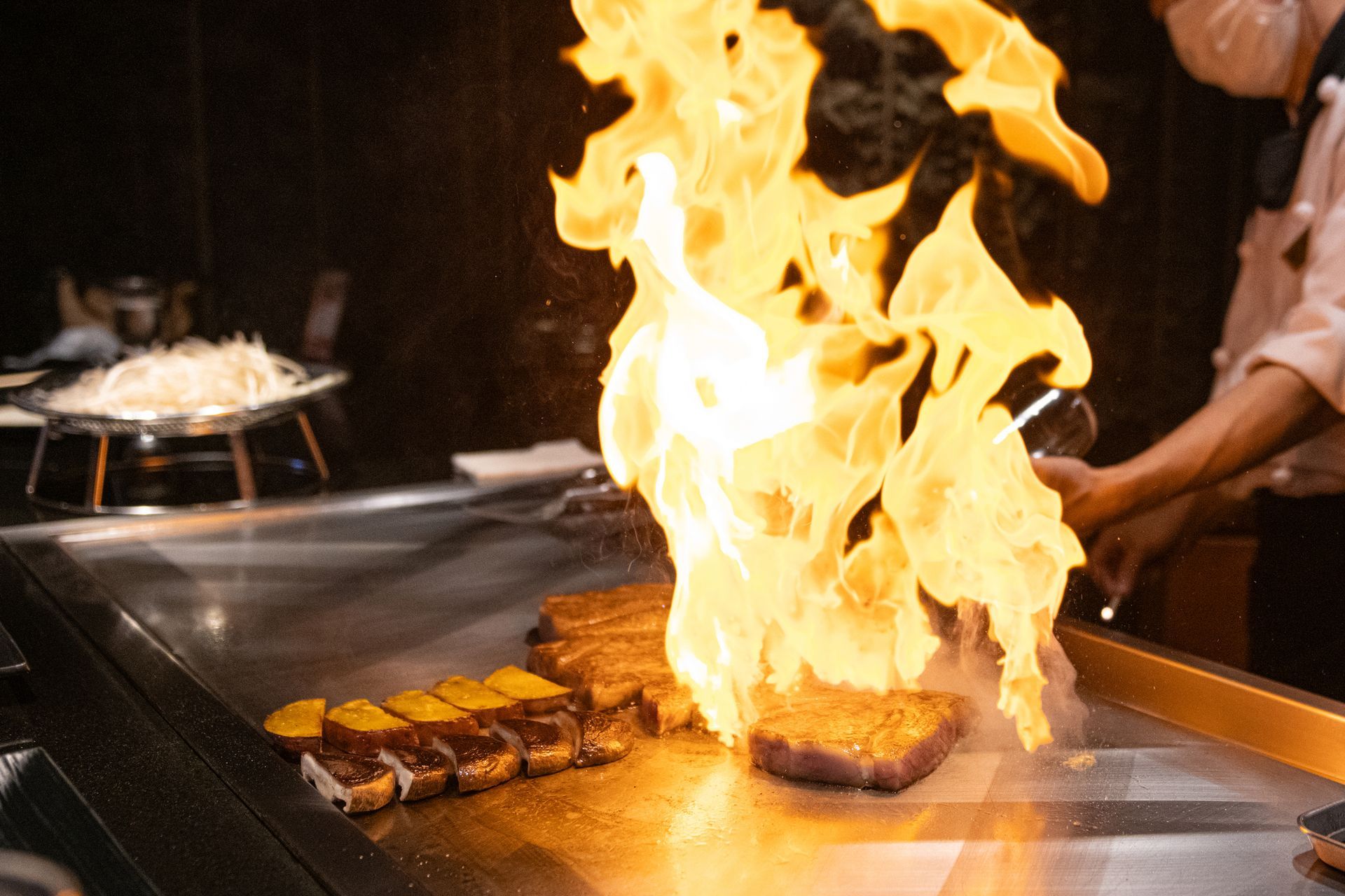 Chef cooking steak on teppanyaki grill with large flames.