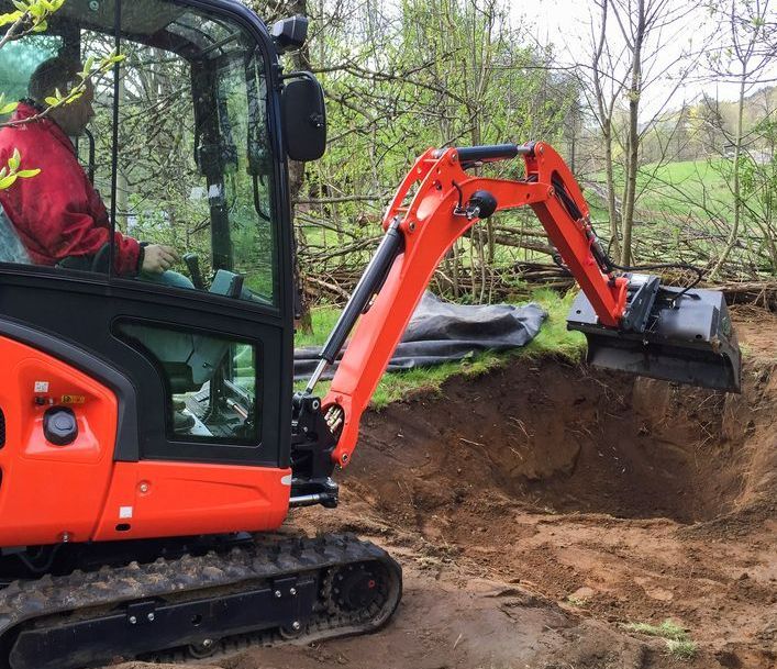 A Man is Driving a Small Orange Excavator in a Dirt Field — Puzzles Plumbing & Excavations In Koonorigan, NSW