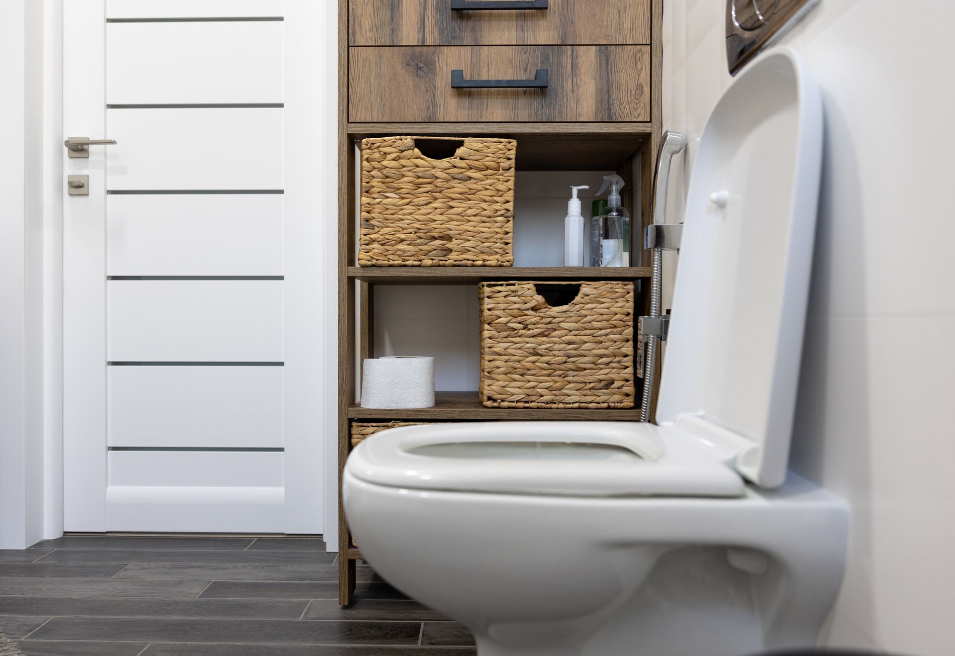 White toilet, open, next to a wooden storage unit with wicker baskets and a white door.