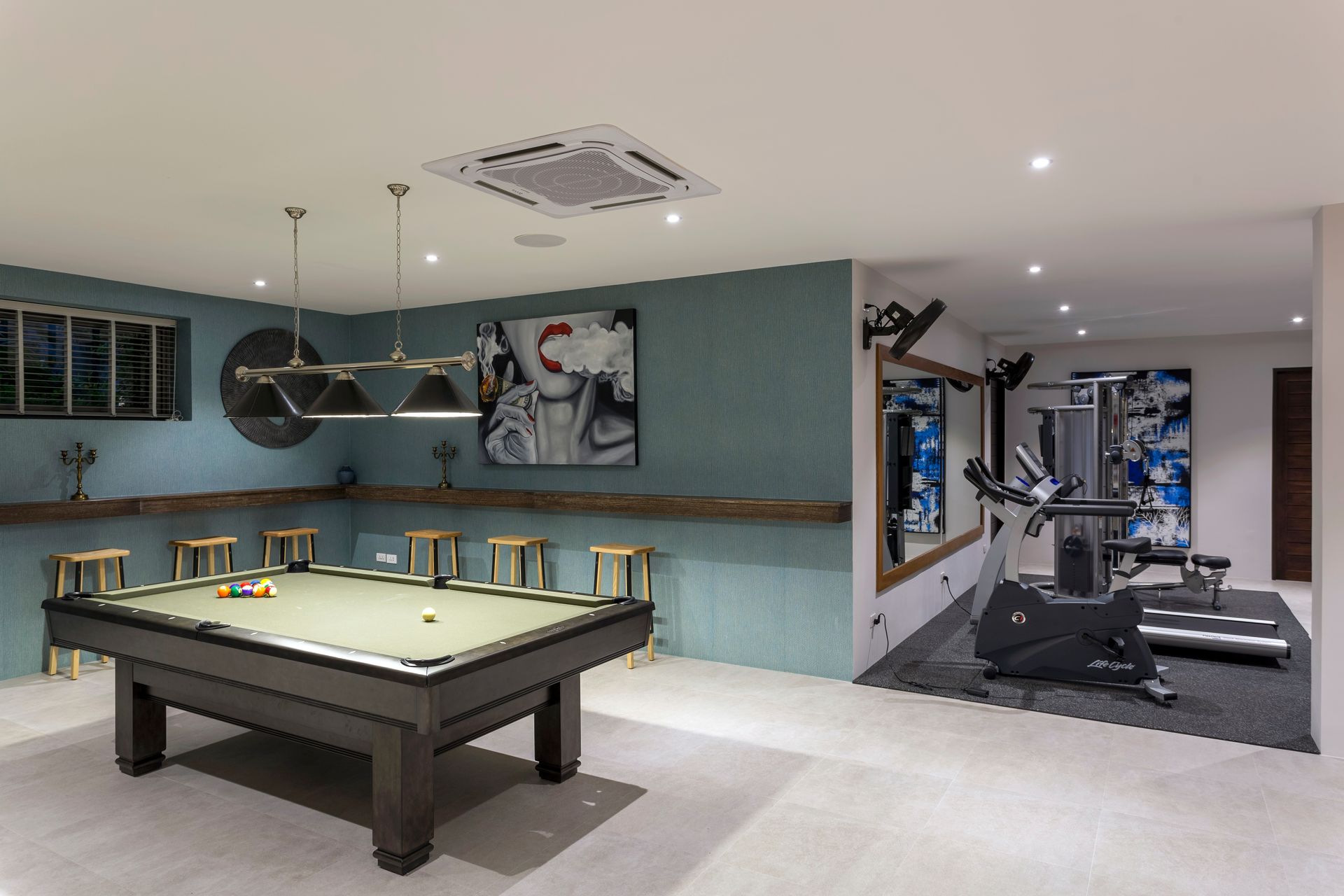 Pool table in a game room with a small gym. Teal walls, stools, and art.