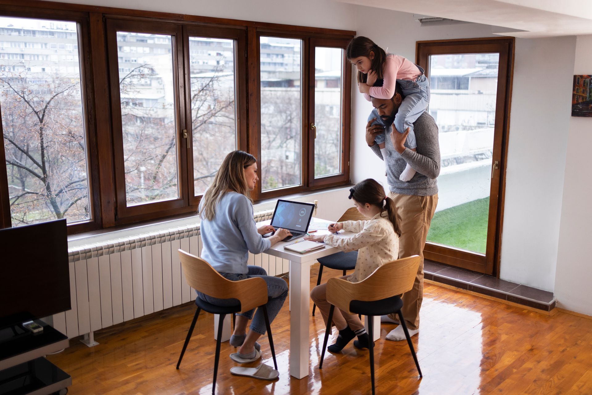 Family in a home, woman working on laptop, man with child on shoulders, another child at table.